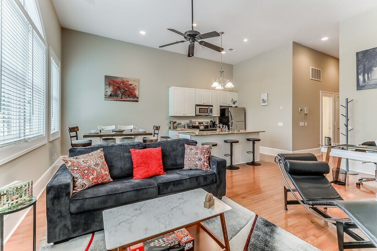 A modern living room with a dark grey sofa, a marble coffee table, and a black reclining chair. A ceiling fan is visible, and there is a kitchen area with white cabinets and a microwave.
