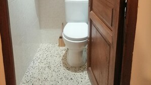 A small bathroom with a white toilet, a wooden shelf above it, and a roll of toilet paper on the wall.