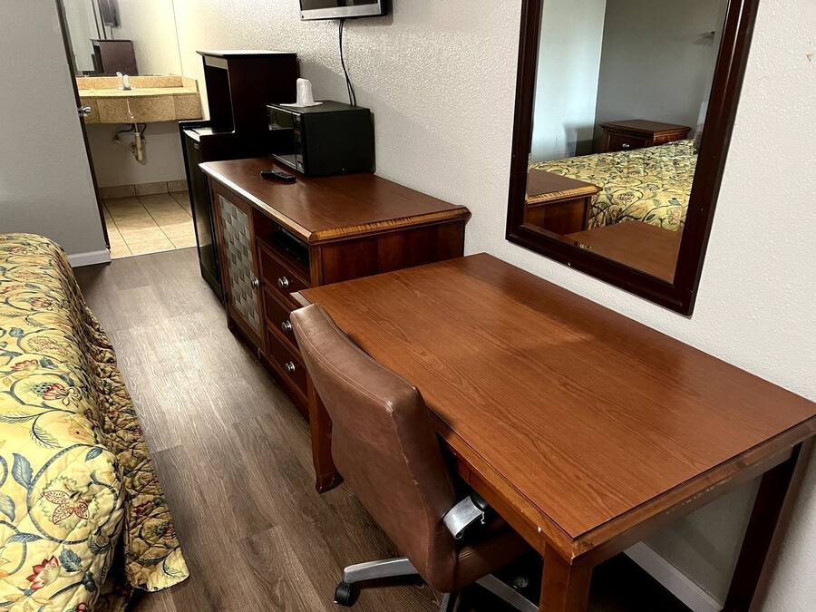 A hotel room with a wooden desk, a chair, a bed with floral bedding, a dresser, a television, and a mirror.