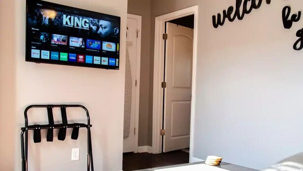 Sala de estar com televisão de tela plana exibindo a interface de um serviço de streaming, um suporte de metal preto, uma porta branca e uma parede com um letreiro de 'bem-vindo'.