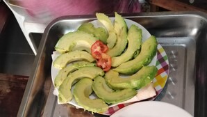 Una bandeja con dos platos de comida, uno con rebanadas de aguacate y un tomate cherry, y el otro con trozos de tortilla.