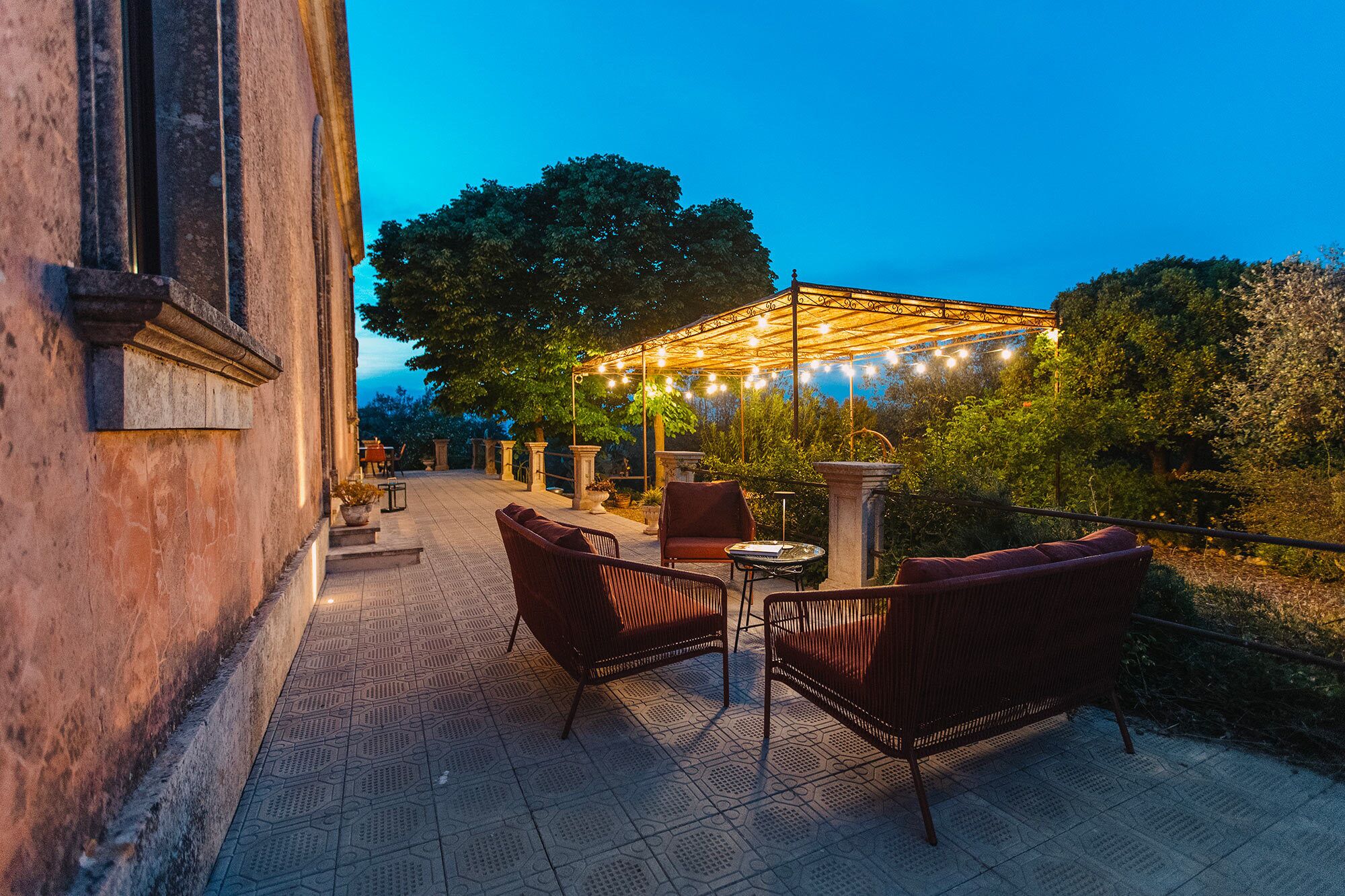 An outdoor patio with wicker furniture, a pergola with string lights, and a stone wall.