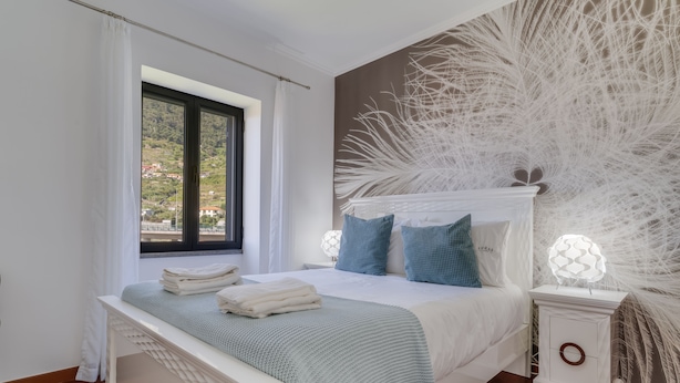 A bedroom with a large bed, a nightstand, a window with curtains, and a wall with a feather-like design. - Villa Paixão (Machico)