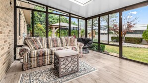Une véranda avec un canapé d’angle, un fauteuil noir et un pouf à motifs.