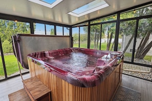 Un bain à remous doté d’une structure extérieure en bois et d’une paroi en verre, entouré d’une terrasse offrant une vue sur un paysage verdoyant.