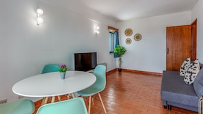 Uma sala de estar moderna com uma mesa redonda, cadeiras verde-água, um sofá cinzento e uma televisão.