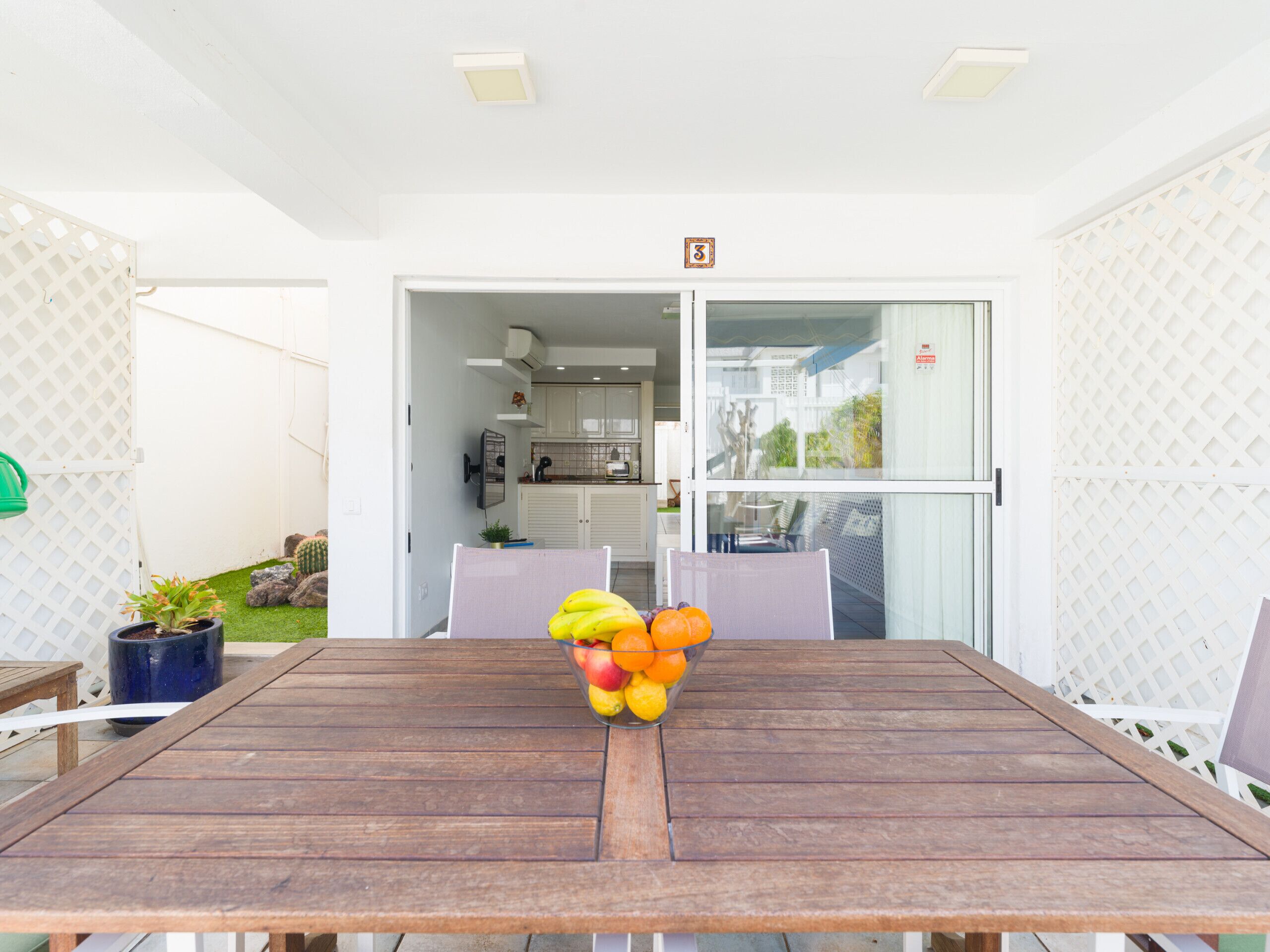 Une table en bois avec un panier de fruits au centre, une plante en pot et une vue sur la cuisine et l’espace patio.