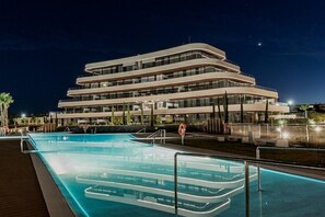 Un hôtel moderne à l’architecture imposante, doté de plusieurs étages, d’une piscine et d’un espace extérieur bien éclairé.