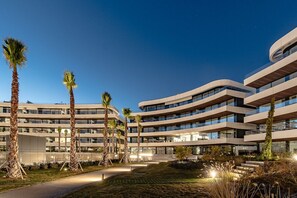 Un bâtiment moderne à plusieurs étages, doté d’une façade incurvée, entouré de palmiers et bordé d’un chemin bien éclairé.