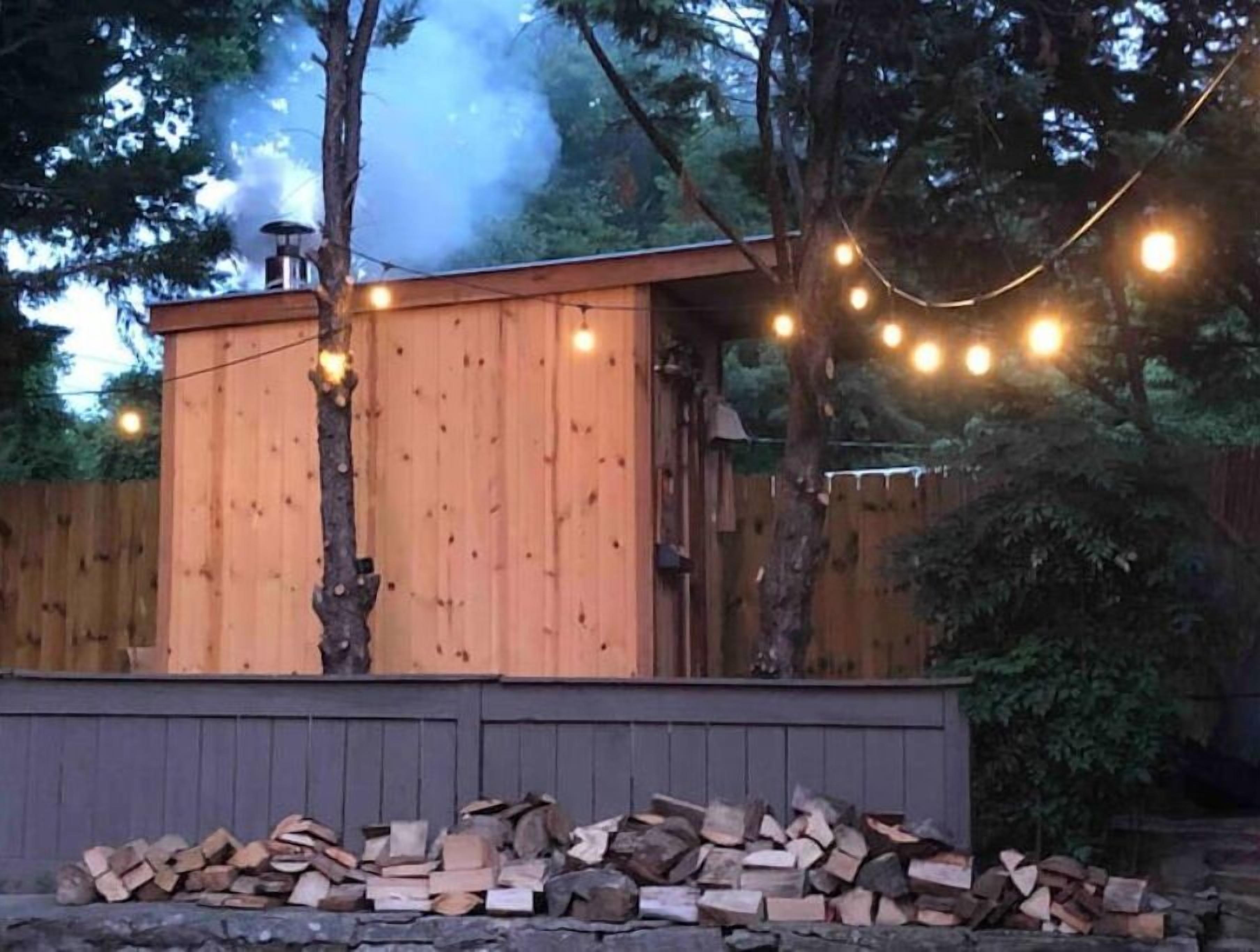 Una cabaña de madera con chimenea humeante, rodeada por una valla de madera y árboles.