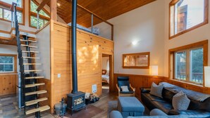 Een gezellig chaletinterieur met een slaapkamer op de vide, een open haard, een trap en een zithoek met een bank en een fauteuil.