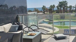 Een balkon met rieten meubilair, een glazen hekwerk en uitzicht op een zwembad en palmbomen.