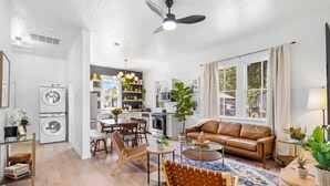A modern living room with a sofa, coffee table, and chairs, a kitchen area with a dining table, and a laundry area with a washing machine.