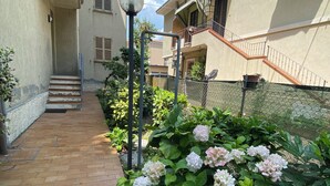 A residential building with a brick pathway leading to a staircase, surrounded by lush greenery and flowering plants.