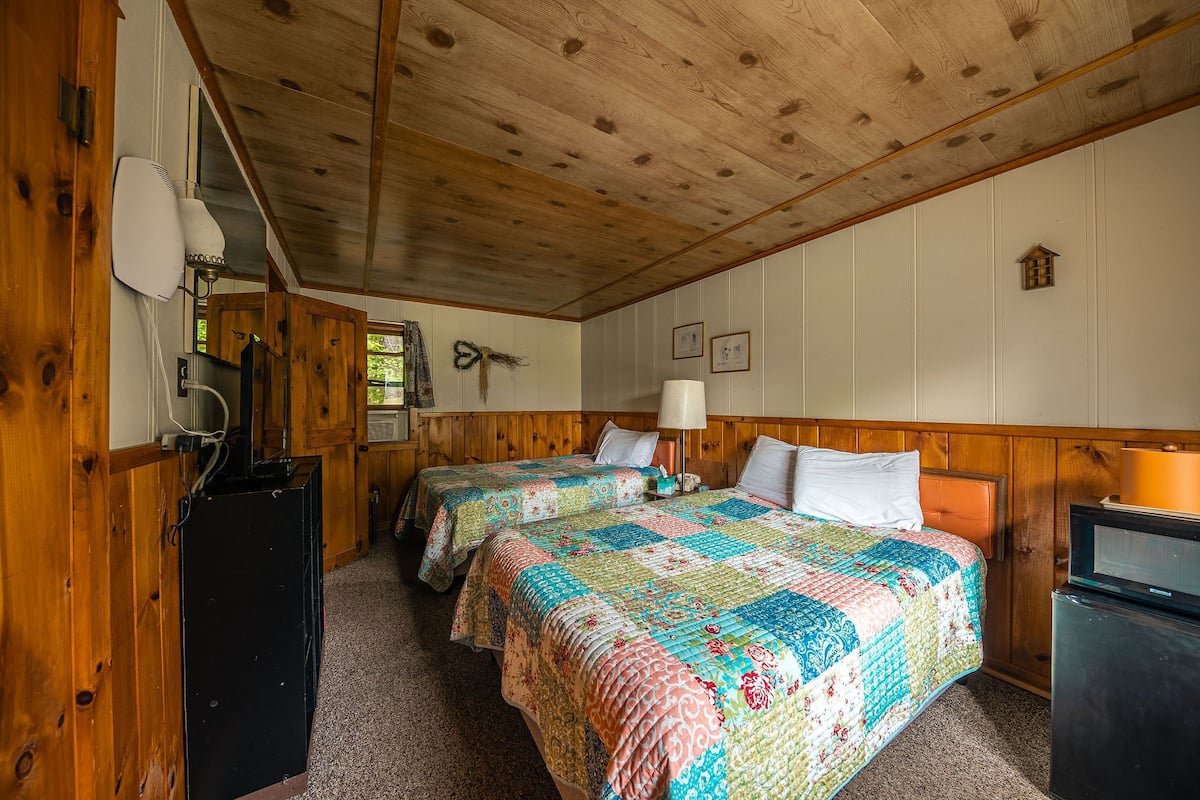Une chambre de style cabane avec deux lits, des murs en bois, un ventilateur de plafond et un petit téléviseur.