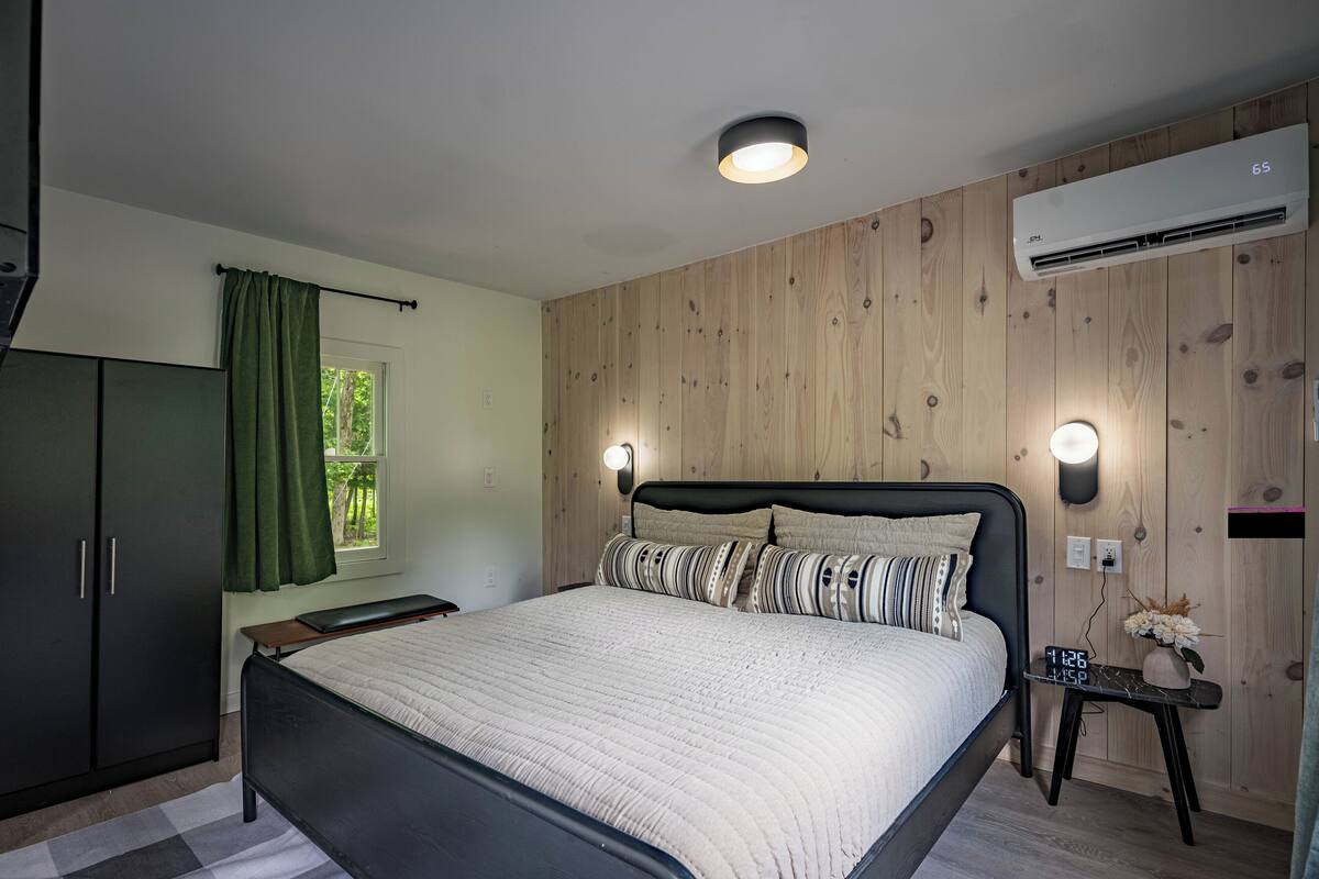 Une chambre à coucher comprenant un lit, une armoire, une fenêtre avec des rideaux, un appareil de climatisation et une table de chevet avec une horloge et des fleurs.