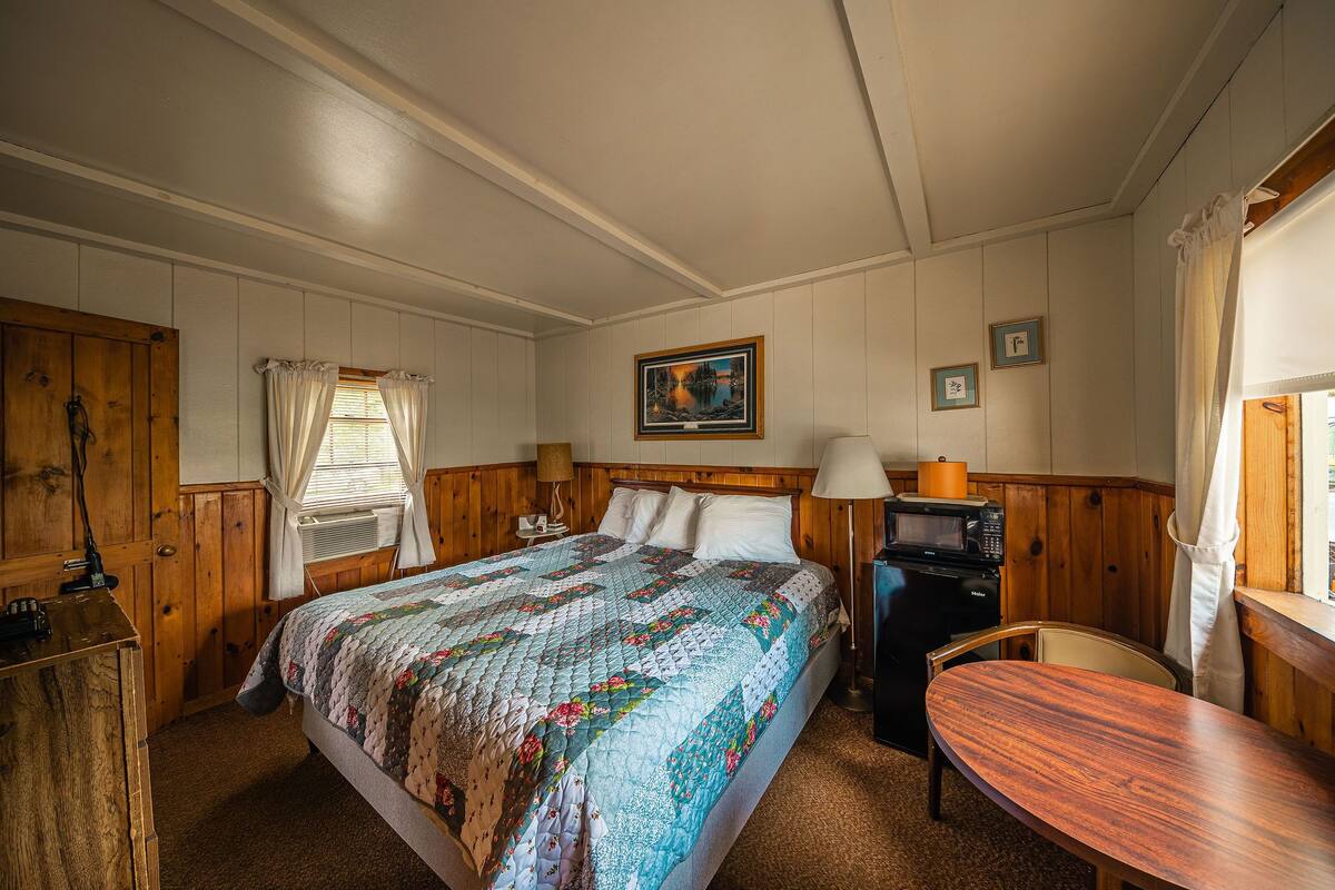 Une chambre avec un lit en bois, un bureau, une chaise, un four à micro-ondes et une fenêtre avec des rideaux.