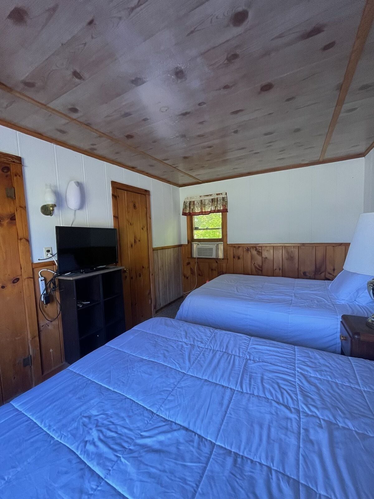A room with two beds, wooden walls, a television, and a window with a curtain.