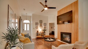 A modern living room with a fireplace, wooden paneling, a ceiling fan, and a flat-screen TV mounted above the fireplace. - Shelby Farms Greenline Access: Modern Cordova Home (Cordova)