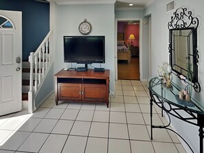 Un couloir avec une console en bois, un téléviseur à écran plat, une horloge et une table en verre ornée d’objets décoratifs.