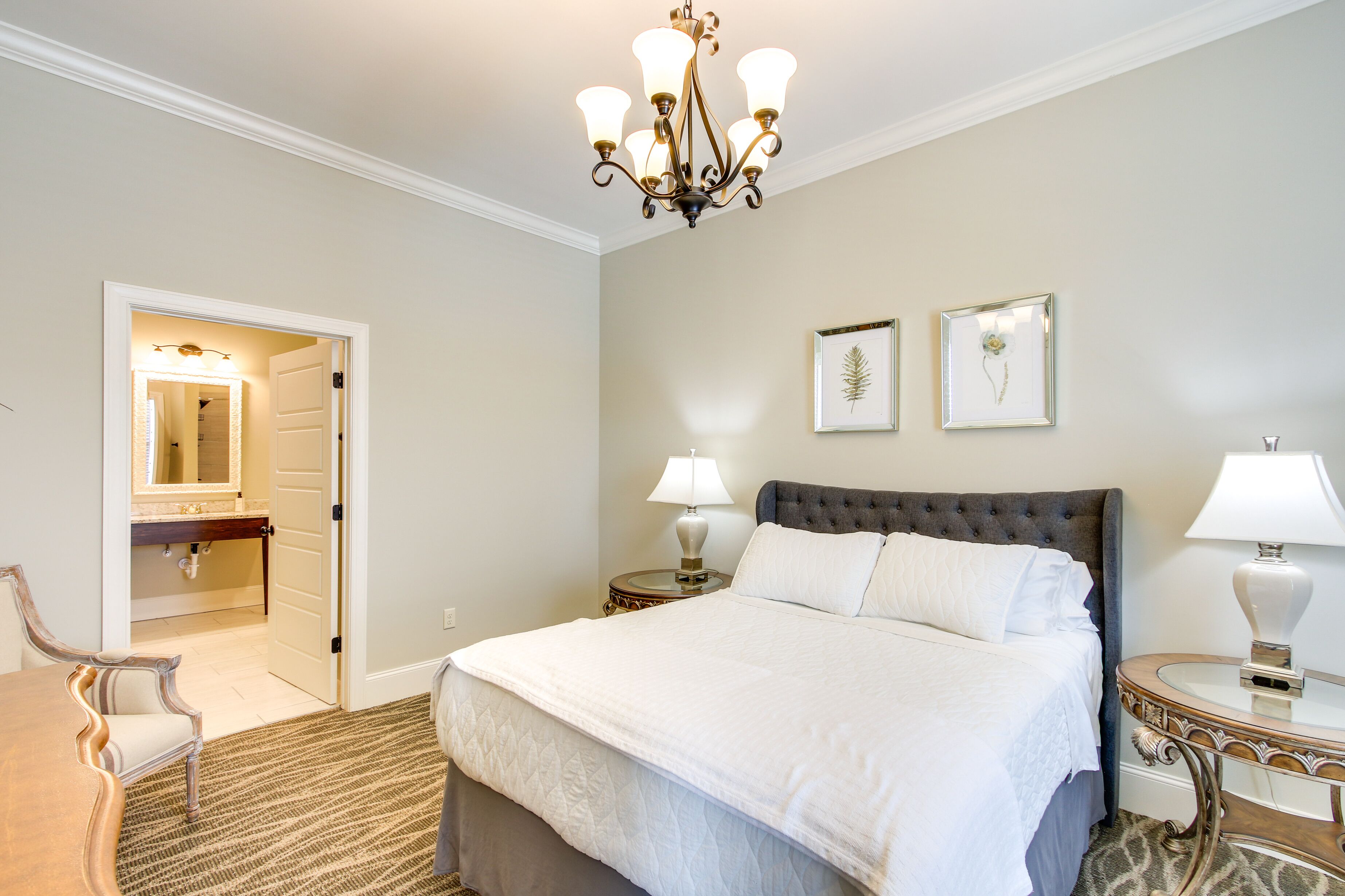 A hotel room with a large bed, two bedside tables with lamps, a chandelier, a mirror, and a door leading to a bathroom.