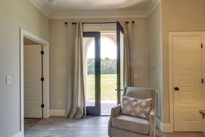 Un pasillo con una puerta que da a un patio, una silla con cojín estampado y vista a un paisaje verde a través de la puerta.