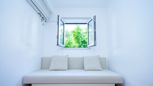 Un sofà blanc amb tres coixins en una habitació de parets blanques i una finestra amb persianes obertes.