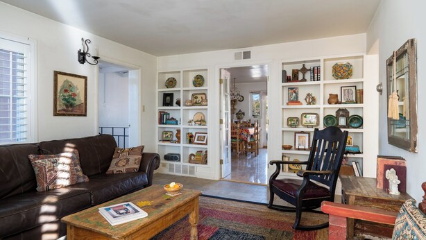 A living room with a brown leather sofa, a wooden coffee table, and a black chair. There is a bookshelf filled with various decorative items and a dining area in the background.