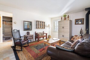 A living room with a brown leather sofa, a wooden rocking chair, a wooden coffee table, and a patterned rug. There is a white cabinet with decorative items on top and a mirror on the wall. - Casita de Jeanetta by Kokopelli Property Management (Santa Fe)
