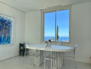 Un comedor moderno con una mesa redonda y sillas transparentes, ventanas amplias con vista al mar y un cuadro abstracto y colorido en la pared.