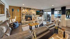 A cozy living room with a stone fireplace, leather sofas, and a view of the ocean. - Relax on the Bay — Kayaks, Hot Tub & Firepit Nights (Ellison Bay)