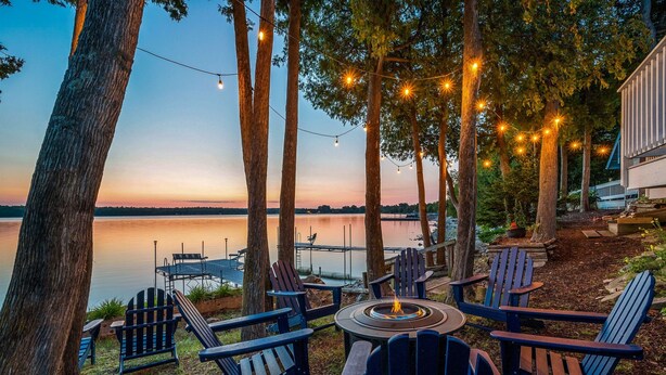A lakeside seating area with Adirondack chairs and a fire pit, surrounded by trees and string lights. - Relax on the Bay — Kayaks, Hot Tub & Firepit Nights (Ellison Bay)