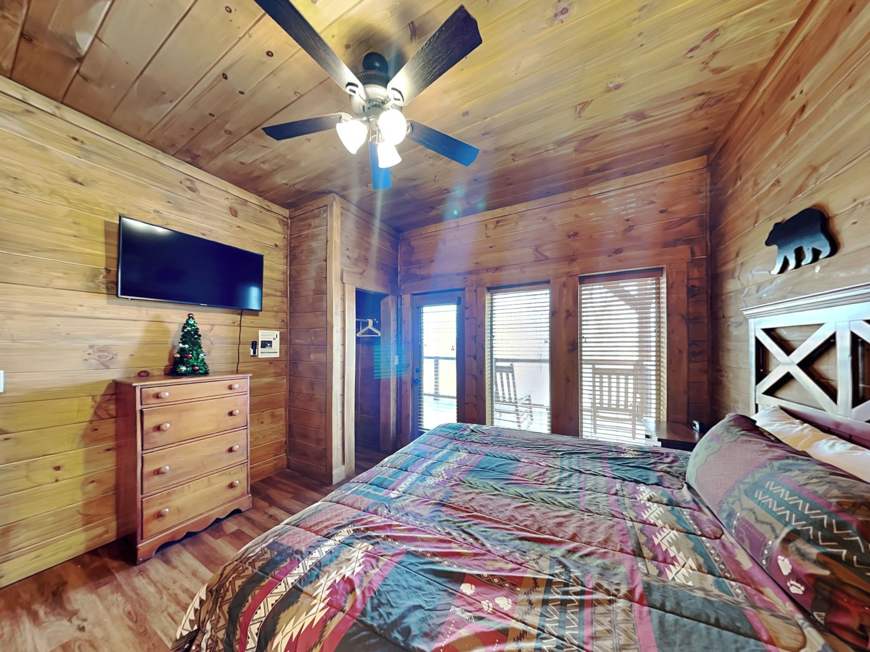 A wooden cabin bedroom with a bed, dresser, TV, and a ceiling fan.