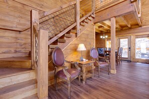 A wooden interior with a staircase, a dining area with chairs and a table, and a view through a window.