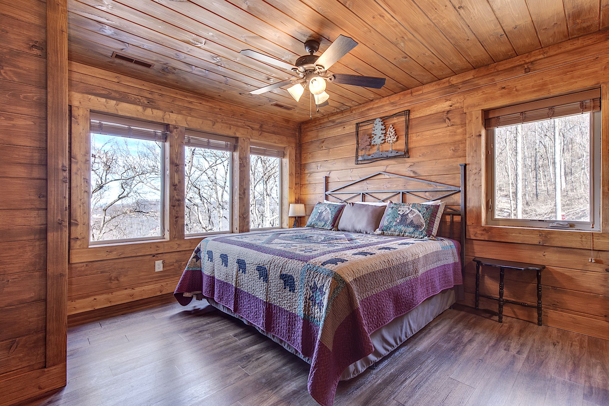Une chambre dans une cabane en bois, avec un lit, un ventilateur de plafond et de grandes fenêtres.
