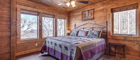 Une chambre dans une cabane en bois, avec un lit, un ventilateur de plafond et de grandes fenêtres.