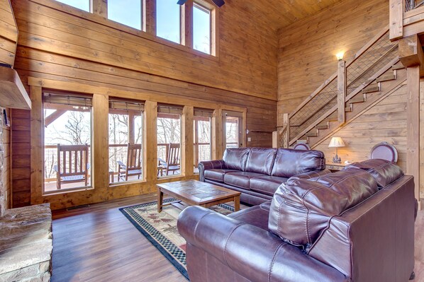 A spacious living room with a large wooden ceiling, a stone fireplace, and a staircase.