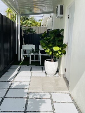 Un patio couvert avec un sol carrelé, une table blanche, une chaise blanche et une grande plante en pot.