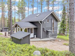 Een chalet met twee verdiepingen, een veranda, een klein tuinhuisje en een metalen trap naar een terras.