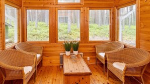 Eine Holzhütte mit großen Fenstern, Korbmöbeln und einem zentralen Tisch mit Topfpflanzen.