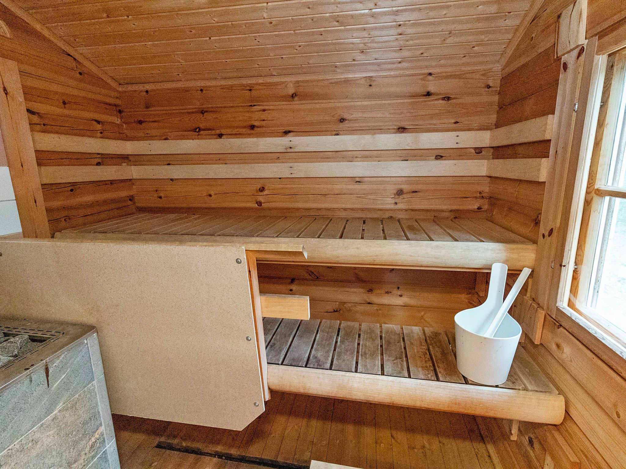 Puinen mökki sisältä, siellä on penkki, valkoinen ämpäri ja ikkuna.