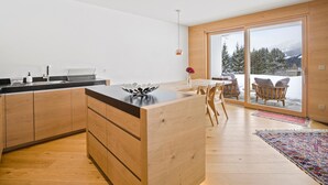 Una cucina moderna con mobili in legno, un piano di lavoro nero e una grande isola. C'è una zona pranzo con tavolo e sedie, e vista su un paesaggio innevato attraverso una porta vetrata.
