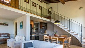 A spacious living area with a sofa, a dining table with chairs, a kitchen in the background, and a staircase leading to an upper level.
