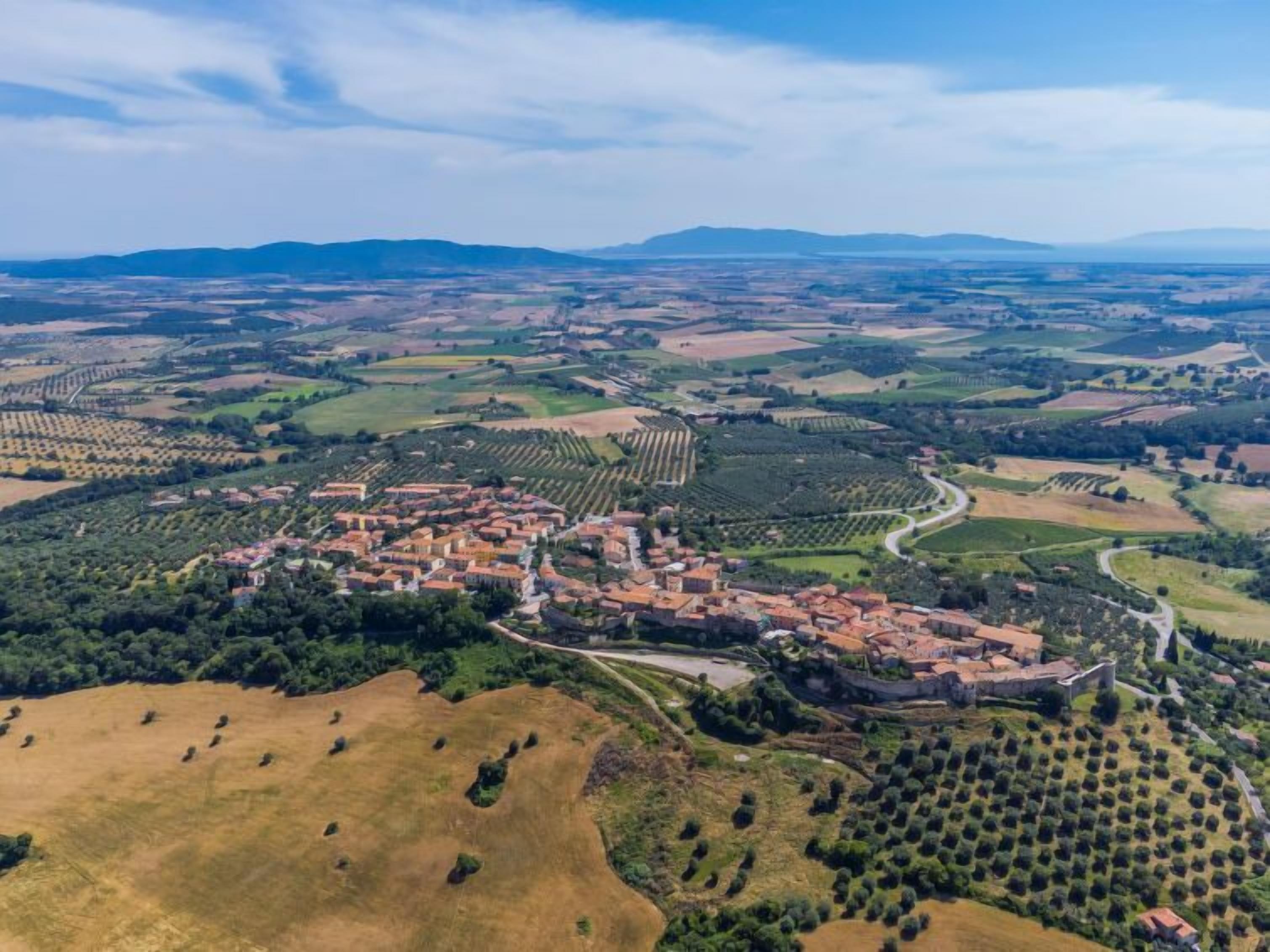 An aerial view of a town with terraced vineyards, a central square with buildings, and rolling hills in the background.
