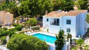 Eine weiße Villa mit Swimmingpool, umgeben von üppiger Vegetation und einer Hügelsicht auf bunte Häuser.
