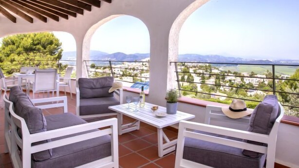 Une terrasse dotée de mobilier blanc, d’une table et offrant une vue sur un paysage composé de bâtiments et de montagnes.