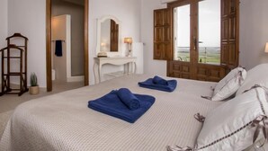 Une chambre d’hôtel avec un grand lit, un bureau avec un miroir, un lustre et une fenêtre avec des volets en bois.