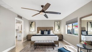 A bedroom with a large bed, a ceiling fan, a nightstand, a bench, a small table with a vase, and a window with a view.