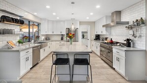 A modern kitchen with white cabinetry, a central island with two chairs, stainless steel appliances, and a variety of kitchen tools and decorations.