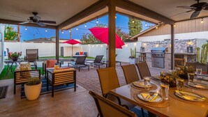 An outdoor patio with a dining table set for four, surrounded by chairs. There are ceiling fans, string lights, and a red umbrella.